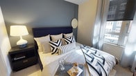 This single-floor residence showcases a thoughtfully designed interior with a modern aesthetic that balances comfort and style. The bedroom serves as a standout feature, centered around a bold slate-blue accent wall that creates a striking focal point behind the upholstered navy headboard with elegant brass stud detailing. The bedding is dressed in a sophisticated monochromatic palette of whites, grays, and deep navy, accented by geometric-patterned pillows and a textured throw that add depth and visual interest. A round wall mirror adds a decorative touch while enhancing the sense of space. Natural light filters through sheer curtains, creating a soft, inviting ambiance throughout the room. The warm beige tones of the surrounding walls complement the cooler accent wall, achieving a balanced and cohesive color scheme. A glass geometric lantern and decorative accessories on the bedside tray contribute to the refined, curated feel of the space. The overall design conveys a contemporary sensibility with careful attention to detail, making this residence a comfortable and visually appealing living environment.