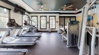 This fitness center is designed to offer a comprehensive workout experience within a thoughtfully appointed single-level space. The layout is organized to accommodate a wide variety of training disciplines, with distinct zones for cardio, strength training, and functional fitness. The cardio area features a row of treadmills, elliptical machines, and stair climbers positioned alongside large, dark-trimmed windows that invite natural light and views of the surrounding greenery, creating an energizing atmosphere during workouts. Ceiling fans overhead contribute to a comfortable environment throughout the space.

The strength training zone is generously stocked with Precor cable machines, a full rack of kettlebells, barbell weight racks, a flat adjustable bench, and an extensive dumbbell selection, all reflected in floor-to-ceiling mirrors that enhance the sense of openness. Rubber flooring spans the entire facility, providing both durability and safety underfoot. A dedicated functional fitness area houses stability balls and step platforms alongside mounted flat-screen televisions for added convenience. Recessed lighting and a dark tray ceiling with crown molding give the space a polished, upscale aesthetic that elevates the overall fitness experience.