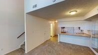 This two-story space is defined by its striking vertical design, most evident in the living area where soaring double-height ceilings create an airy, expansive atmosphere. Large wall-mounted mirrors along one side of the living area amplify the sense of space, while carpet flooring in warm neutral tones runs throughout the main living zones. A staircase with a wood handrail connects the two levels, anchored beneath an open upper-level overhang that adds architectural dimension to the lower floor.

The kitchen is well-appointed with rich, dark wood cabinetry, a double-basin sink, a dishwasher, and a tile backsplash in soft blue-green tones. A peninsula with a laminate countertop provides a natural pass-through between the kitchen and the living area. Tile flooring and a flush-mounted fluorescent light fixture complete the functional space.

Upstairs, the bedroom offers a ceiling fan with a clerestory window for natural light, and a textured ceiling. A set of bifold closet doors provides ample storage, while an adjoining bathroom features white cabinetry, a vanity with a well-lit mirror, and a tub-shower combination with smooth surround panels.