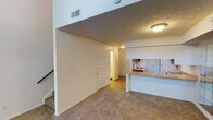 This two-story space is defined by its striking vertical design, most evident in the living area where soaring double-height ceilings create an airy, expansive atmosphere. Large wall-mounted mirrors along one side of the living area amplify the sense of space, while carpet flooring in warm neutral tones runs throughout the main living zones. A staircase with a wood handrail connects the two levels, anchored beneath an open upper-level overhang that adds architectural dimension to the lower floor.

The kitchen is well-appointed with rich, dark wood cabinetry, a double-basin sink, a dishwasher, and a tile backsplash in soft blue-green tones. A peninsula with a laminate countertop provides a natural pass-through between the kitchen and the living area. Tile flooring and a flush-mounted fluorescent light fixture complete the functional space.

Upstairs, the bedroom offers a ceiling fan with a clerestory window for natural light, and a textured ceiling. A set of bifold closet doors provides ample storage, while an adjoining bathroom features white cabinetry, a vanity with a well-lit mirror, and a tub-shower combination with smooth surround panels.