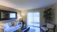 This single-floor space showcases a well-appointed living area designed with both comfort and functionality in mind. The room features warm, neutral-toned walls that create an inviting atmosphere, complemented by smooth, light-colored ceilings that enhance the sense of openness. A large sliding glass door dressed with vertical blinds serves as a prominent architectural feature, allowing natural light to filter into the space while providing direct access to an outdoor area. The living area is thoughtfully arranged with a plush sofa adorned with accent pillows, a tufted leather ottoman that doubles as a coffee table, and an additional seating chair. A decorative area rug anchors the seating arrangement atop hardwood-style flooring, adding texture and warmth to the space. A stylish console table along the wall holds a table lamp and decorative botanicals, providing ambient lighting that complements the natural light. A bold, framed mirror-like wall accent adds a modern touch to the décor. The overall ambiance is relaxed yet polished, making it a comfortable and visually cohesive living environment.