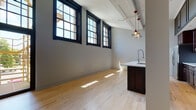 Located at 215 N Webster Ave in Green Bay, Wisconsin, this thoughtfully designed single-floor unit blends industrial character with modern finishes to create a stylish and functional living space. The open-concept layout is anchored by warm light maple hardwood flooring that runs throughout, complemented by soft gray walls and exposed structural ceiling elements that nod to the building's architectural heritage.

Large, black-framed multi-pane windows line the main living area, flooding the space with abundant natural light and offering views of the surrounding brick exterior and outdoor stairway access. The expansive open living area flows seamlessly into the kitchen, which serves as a true centerpiece of the unit. Rich dark-stained cabinetry — extending to the ceiling with upper storage — pairs elegantly with a white quartz waterfall island, stainless steel appliances including a French door refrigerator and range, a subway tile backsplash, and a striking industrial-style pendant lighting duo overhead.

The bathroom is finished with classic white wall tile, a glass-enclosed walk-in shower with dark hardware accents, and distinctive black penny tile flooring, adding a refined yet edgy touch. In-unit laundry is also present. The unit includes access to an outdoor deck area, enhancing the overall livability of this urban retreat.