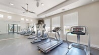 Located at 1891 N Litchfield Rd in Goodyear, Arizona, this fitness center offers a well-appointed exercise space designed to support a full range of workout routines. The single-floor layout provides ample open floor space, finished with durable rubber flooring ideal for both cardio and strength training activities.

The room is bathed in natural light through multiple windows fitted with white blinds, offering views of the surrounding community, including glimpses of a pool area. A tray ceiling adds architectural interest overhead, while multiple ceiling fans ensure comfortable air circulation throughout the space. Recessed lighting provides consistent illumination for all areas of the room.

The fitness center is equipped with a comprehensive selection of exercise equipment, including treadmills, an elliptical trainer, a stationary bike, and a multi-station cable strength machine. A full dumbbell rack provides a range of free weight options, complemented by an adjustable workout bench. Floor-to-ceiling mirrors line one wall, enhancing the sense of openness and allowing users to monitor their form during exercise.

Glass double doors connect the fitness center to an adjacent common area, adding to the open, accessible feel of the space. The clean, neutral color palette of soft gray and white walls creates a modern, motivating environment well-suited for daily fitness use.