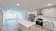Located at 15120 Idlewild Road in Matthews, NC, this well-appointed one-bedroom, one-bathroom residence offers a thoughtfully designed single-floor layout with a clean, modern aesthetic throughout. The open-concept living and kitchen area is bright and airy, featuring light wood-look flooring, recessed lighting, and large windows that invite abundant natural light. A glass-paneled door leads to a private balcony overlooking lush greenery and mature trees, providing a peaceful outdoor retreat. The kitchen is a standout, showcasing gray shaker-style cabinetry with matte black hardware, crisp white quartz countertops, a classic subway tile backsplash, stainless steel appliances including a French door refrigerator and range, a wall-mounted range hood, and a central island perfect for dining or meal prep. The spacious bedroom is finished with plush carpet, a ceiling fan, and a wide window framing outdoor views. It connects directly to a walk-in closet with built-in wire shelving and to the full bathroom, which features a white vanity, wood-look tile flooring, and a tiled walk-in shower with a sleek black-framed glass enclosure. Clean white walls and consistent modern finishes tie the entire space together cohesively.