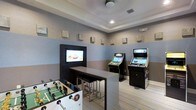 This single-floor game room is thoughtfully designed as a social and recreational hub. The space features a distinctive two-tone blue and gray color scheme along the walls, accented by decorative stone block wall art that adds a textural, contemporary touch. A tray ceiling with recessed lighting contributes to a polished, finished aesthetic overhead, while light wood-toned flooring adds warmth underfoot.

At the heart of the room, a wall-mounted flat-screen television is paired with a bar-height counter and stools, creating an ideal spot for viewing and socializing. Two upright arcade gaming machines are positioned nearby, offering interactive entertainment options. A foosball table occupies the foreground, providing a classic, engaging game for multiple players.

The layout is open and functional, allowing easy movement between the various entertainment stations. Subtle wall-mounted speakers suggest an integrated audio experience throughout the room. The overall ambiance strikes a balance between a casual lounge and an active game room, making it well-suited for group gatherings, leisure activities, and social interaction across all age groups.