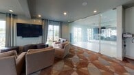 This single-floor community amenity space in Henrico, Virginia, is thoughtfully designed to accommodate a variety of recreational and social activities. The interior is unified by a distinctive geometric-patterned carpet in warm earth tones of brown, tan, and gray, which flows throughout the space and contributes to its cohesive, contemporary aesthetic. Recessed lighting and pendant fixtures illuminate the rooms, complemented by generous natural light streaming through large floor-to-ceiling windows that frame views of the surrounding neighborhood.

The lounge area features comfortable upholstered seating arranged around a wall-mounted flat-screen television, creating an inviting setting for casual gatherings. Adjacent to the lounge, a dedicated game room houses a full-sized billiards table beneath an elegant chandelier, alongside additional wall-mounted display screens. A separate card and game room offers multiple round tables with cushioned chairs, accented by contemporary artwork and direct access to an outdoor balcony. A glass-partitioned fitness or activity studio with mirrored walls and ceiling fans is also visible within the space. Accessible restroom facilities are conveniently located nearby, rounding out this well-appointed amenity area.