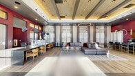 Located at 12479 Rojas Dr in Horizon City, Texas, this single-floor community clubhouse offers a spacious, open-plan interior designed with contemporary style and high-end finishes throughout. The space is defined by its striking coffered ceiling with warm wood-tone paneling and recessed lighting, complemented by elegant circular pendant fixtures that add a modern, sculptural quality to the overhead design. Tall windows dressed with sheer drapes and plantation shutters line the perimeter, flooding the interior with abundant natural light and creating an airy, welcoming ambiance. The flooring transitions seamlessly between large-format tile and wood-look planks, reinforcing the space's refined aesthetic. A generously sized reception and service counter with a sleek stone surface serves as a central focal point, accented by sculptural ring pendant lights. Adjacent to it, a bar-height counter with warm-toned seating provides a casual gathering area. The lounge zone features a large sectional sofa with coordinating accent pillows in burgundy and blush, arranged around a coffee table atop a textured area rug. Along the windowed wall, bistro-style tables and upholstered chairs offer additional seating. Bold burgundy accent walls and vibrant artwork near the entryway create a dynamic visual contrast, while glass-partitioned office areas provide functional workspace within the overall layout.