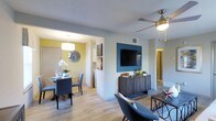 This single-floor residence features an open-concept layout that seamlessly connects the living and dining areas, creating a cohesive and functional living space. The living area is anchored by a teal accent wall that frames a wall-mounted flat-screen television, complemented by a rustic wood media console below. A ceiling fan with integrated lighting adds both practicality and style overhead. Light wood-tone flooring runs throughout, brightening the space and lending a clean, modern aesthetic. The dining area is distinguished by a bold yellow accent wall that adds a vibrant pop of color, and is furnished with a round dining table surrounded by upholstered gray chairs. A contemporary pendant light hangs above the table, while a round decorative mirror on the wall enhances the sense of depth. The two spaces are separated by a partial dividing wall, maintaining an open feel while defining each zone. A hallway visible in the background suggests additional rooms beyond. The overall ambiance is modern and cheerful, with a thoughtful use of contrasting accent colors, consistent flooring, and layered lighting that creates a warm and inviting atmosphere throughout.
