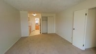 This single-floor space offers a straightforward and functional layout designed for comfortable everyday living. The main room is finished with wall-to-wall carpet in a neutral beige tone, creating a warm and understated atmosphere throughout. A textured popcorn ceiling adds a classic touch overhead, while the walls are painted in a soft, light cream that keeps the space feeling open and bright.

A built-in closet with sliding doors provides convenient storage directly within the main room, offering practical organization without occupying additional floor space. The doors are finished in white, blending seamlessly with the surrounding walls.

An open passageway connects the main room to the kitchen, which is finished with tile or linoleum flooring for easy maintenance. The kitchen features warm wood-toned cabinetry along the lower and upper walls, providing ample storage. A white refrigerator and additional appliances are visible, along with a countertop workspace. A window above the sink area allows natural light to filter into the kitchen, brightening the functional workspace. Overall, the space presents a clean, no-frills environment with practical amenities throughout.