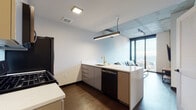 This contemporary urban unit is thoughtfully designed with a cohesive modern aesthetic throughout. The open-concept kitchen and living area serve as the heart of the space, where dark hardwood flooring grounds the layout and exposed concrete ceiling elements add an industrial character. The kitchen is outfitted with sleek, high-gloss grey cabinetry, white quartz countertops, and a suite of black stainless steel appliances including a gas range, microwave, refrigerator, and dishwasher. A gold-toned faucet at the peninsula sink adds a refined accent, while a linear pendant light overhead enhances the workspace.

The living area transitions seamlessly from the kitchen and is anchored by floor-to-ceiling windows that frame sweeping city views, flooding the space with natural light. The bedroom continues the floor-to-ceiling window design, offering expansive urban vistas that make for a striking backdrop. Exposed concrete columns punctuate the room, reinforcing the building's industrial-modern architecture.

The bathroom is finished with large-format grey tile in a glass-enclosed walk-in shower, complemented by matte black fixtures and hardware throughout, delivering a clean, upscale finish. Every detail of this space reflects a consistent design language that balances modern minimalism with urban sophistication.