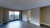 This single-floor space features a bright, open living area finished with smooth hardwood-style flooring in a cool grey tone, lending a clean and modern aesthetic throughout. The walls are painted in a neutral greige hue, complemented by crisp white crown molding and baseboards that frame the room with a polished finish. A textured ceiling adds subtle visual interest overhead.

A large sliding glass door dominates one wall, flooding the space with natural light and providing direct access to an outdoor balcony, which overlooks what appears to be a neighboring structure with a distinctive green awning. The balcony extends the living area outdoors and adds an appealing layer of functionality to the space.

A six-panel interior door opens to an adjoining room, partially visible and appearing to contain tiled flooring, suggesting a bathroom or utility space. A wall-mounted heating unit is positioned beneath the sliding door, ensuring practical climate control.

The overall ambiance is clean, neutral, and move-in ready, offering a versatile blank canvas that accommodates a range of interior styles and furnishing preferences.