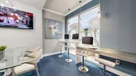 This thoughtfully designed single-floor amenity space offers a cohesive and contemporary environment that blends social lounging with productive work areas. The space is anchored by a striking lounge zone featuring a bold, textured blue wave-panel accent wall, plush royal blue velvet armchairs, and glass-topped coffee tables that create an inviting yet sophisticated atmosphere. Patterned carpet tiles in varying shades of blue and gray define each functional area with visual interest underfoot.

A floor-to-ceiling glass partition wall elegantly separates the lounge from an adjacent area, maintaining an open, airy feel while providing a sense of definition. The business center section is lined with sleek, wall-mounted white countertops fitted with desktop computers, illuminated by pendant lights and framed by large windows that flood the area with natural light and offer outdoor views.

Additional features include a wet bar or kitchenette with dark tile backsplash and gray cabinetry, wall-mounted flat-screen televisions, and curated artwork throughout. Crown molding and recessed lighting contribute to a polished architectural finish. The overall ambiance is modern and vibrant, combining practical amenities with upscale design elements.