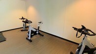 This single-floor fitness space offers a well-organized and functional environment for exercise and physical wellness. The room features a clean, contemporary aesthetic with smooth, light-toned walls and dark baseboards that provide a sharp, polished contrast. The flooring consists of durable, wood-look material well-suited for high-traffic workout use.

The space is outfitted with stationary cycling bikes positioned to allow ample room for movement and use, making it suitable for individual or group cycling sessions. Additional exercise equipment is also present, indicating a versatile setup designed to accommodate a range of fitness activities beyond cycling alone.

Recessed and ambient overhead lighting provides consistent, warm illumination throughout the room, ensuring a comfortable and well-lit workout environment at any time of day. The walls are kept clear and unadorned, maximizing the sense of open space and allowing for flexible equipment arrangement as needs evolve.

Overall, the fitness facility presents a practical and inviting atmosphere with a focus on functionality. Its straightforward layout, durable finishes, and dedicated equipment make it a capable and purpose-built space for regular physical activity.