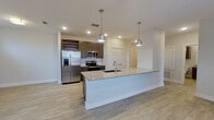 This single-floor space showcases a clean, contemporary design with an open layout that seamlessly connects the kitchen and living areas. Light wood-look flooring runs throughout the main living spaces, complementing the neutral white and gray walls that lend a bright, airy feel to the interior.

The kitchen serves as the focal point of the space, centered around a generous island with a granite countertop, an undermount sink, and pendant lighting overhead. Dark espresso cabinetry provides strong contrast against the white subway tile backsplash, while a full suite of stainless steel appliances—including a side-by-side refrigerator with ice and water dispenser, electric range, over-the-range microwave, and dishwasher—adds a polished, modern touch. A built-in wine rack integrated into the upper cabinetry offers a distinctive storage feature.

A hallway connects the main living area to the private spaces, where soft carpeting transitions into the bedroom area. The bathroom features a glass-enclosed walk-in shower with tiled surround, wood-toned wall cabinetry, and light flooring consistent with the rest of the unit. Recessed lighting throughout provides consistent illumination, enhancing the overall clean and functional ambiance of the space.