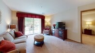This single-floor residence offers a thoughtfully designed layout that balances comfort and functionality across several distinct living areas. The living room is carpeted and welcoming, centered around a media console and accented by a tufted ottoman and a patterned accent chair. Large sliding glass doors flood the space with natural light and provide direct access to an outdoor patio, where views of lush greenery create a serene backdrop. A decorative sunburst mirror and warm lamp lighting add character to the room.

Adjacent to the living room, the dining area features rich hardwood-style flooring, a glass-top dining table with upholstered chairs, and a pendant light overhead. Framed artwork in warm earth tones lines the walls, contributing to the space's cohesive aesthetic.

The kitchen is efficiently arranged in an L-shape configuration, offering ample cabinetry in warm oak tones, white appliances including a refrigerator and dishwasher, and hardwood-style flooring that extends throughout.

The bedroom is a calm retreat with plush carpet, a ceiling fan, and warm ambient lighting. A dedicated workspace adds practical versatility to the room.

Two bathrooms serve the residence — one with a tub-shower combination and one with a walk-in shower — both finished with tile flooring, wood-toned vanities, and soft sage-colored walls.