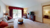 This single-floor residence offers a thoughtfully designed layout that balances comfort and functionality across several distinct living areas. The living room is carpeted and welcoming, centered around a media console and accented by a tufted ottoman and a patterned accent chair. Large sliding glass doors flood the space with natural light and provide direct access to an outdoor patio, where views of lush greenery create a serene backdrop. A decorative sunburst mirror and warm lamp lighting add character to the room.

Adjacent to the living room, the dining area features rich hardwood-style flooring, a glass-top dining table with upholstered chairs, and a pendant light overhead. Framed artwork in warm earth tones lines the walls, contributing to the space's cohesive aesthetic.

The kitchen is efficiently arranged in an L-shape configuration, offering ample cabinetry in warm oak tones, white appliances including a refrigerator and dishwasher, and hardwood-style flooring that extends throughout.

The bedroom is a calm retreat with plush carpet, a ceiling fan, and warm ambient lighting. A dedicated workspace adds practical versatility to the room.

Two bathrooms serve the residence — one with a tub-shower combination and one with a walk-in shower — both finished with tile flooring, wood-toned vanities, and soft sage-colored walls.