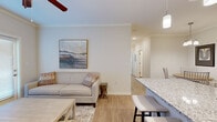 This thoughtfully designed single-floor residence offers three bedrooms and two bathrooms within a compact yet efficient layout. Light wood-style flooring flows throughout the main living areas, creating a cohesive and airy atmosphere complemented by neutral wall tones and crown molding details.

The open-concept living and dining area forms the heart of the home. The living room features a ceiling fan, large windows with access to an exterior door that invites natural light, and ample space for comfortable seating. Adjacent to it, the dedicated dining area accommodates a round table with seating for several guests and is accented by a modern multi-bulb chandelier.

The kitchen is well-appointed with rich dark-brown cabinetry, granite-style countertops, and a full suite of stainless steel appliances including a refrigerator, range, and over-the-range microwave. A breakfast bar provides additional seating and connects seamlessly to the dining space.

The bedrooms are carpeted for comfort, with the secondary bedrooms featuring closets and natural light from windows. The primary bedroom includes a spacious walk-in closet with wire shelving. Both bathrooms are finished with granite-topped vanities, dark cabinetry with chrome hardware, and wood-style flooring. One bathroom features a tub-shower combination, while the other offers a standalone vanity setup.