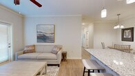 This thoughtfully designed single-floor residence offers three bedrooms and two bathrooms within a compact yet efficient layout. Light wood-style flooring flows throughout the main living areas, creating a cohesive and airy atmosphere complemented by neutral wall tones and crown molding details.

The open-concept living and dining area forms the heart of the home. The living room features a ceiling fan, large windows with access to an exterior door that invites natural light, and ample space for comfortable seating. Adjacent to it, the dedicated dining area accommodates a round table with seating for several guests and is accented by a modern multi-bulb chandelier.

The kitchen is well-appointed with rich dark-brown cabinetry, granite-style countertops, and a full suite of stainless steel appliances including a refrigerator, range, and over-the-range microwave. A breakfast bar provides additional seating and connects seamlessly to the dining space.

The bedrooms are carpeted for comfort, with the secondary bedrooms featuring closets and natural light from windows. The primary bedroom includes a spacious walk-in closet with wire shelving. Both bathrooms are finished with granite-topped vanities, dark cabinetry with chrome hardware, and wood-style flooring. One bathroom features a tub-shower combination, while the other offers a standalone vanity setup.