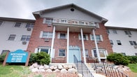 This multi-story brick building presents a timeless and well-maintained exterior that combines traditional architectural elements with a welcoming aesthetic. The facade is constructed of classic red brick, giving the structure a sturdy and enduring character. A prominent columned portico anchors the main entrance, featuring tall white columns that support a decorative upper balcony with a distinctive cross-pattern balustrade. The entrance is framed by a charming light blue double door, adding a touch of color and warmth to the otherwise neutral exterior palette. Above the portico, a circular window detail adds a refined architectural accent beneath the peaked gable, which is finished with light gray lap siding. The building features multiple windows on each level, including units with window air conditioning, indicating individual climate control throughout. A set of steps with black iron railings leads up to the main entrance, with an accessible ramp on one side providing convenient entry for all visitors. The landscaping along the front features natural boulder arrangements interspersed with low shrubs, lending a grounded, rustic quality to the grounds. Overall, the exterior conveys a sense of classic institutional design with residential warmth.
