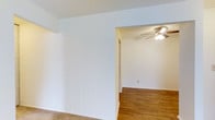 This single-story space offers a straightforward, functional layout with a bright and neutral aesthetic. The interior is finished with crisp white walls throughout, creating a clean and airy atmosphere that allows for versatile decorating possibilities. The flooring transitions between plush carpet in one area and warm hardwood-style planks in the adjoining room, adding visual contrast and defining the separate zones within the space. An open archway connects the two areas, contributing to a sense of flow and continuity. The secondary room is equipped with a ceiling fan fitted with a light fixture, providing both air circulation and ambient lighting. Electrical outlets are positioned along the baseboards, ensuring practical accessibility. The overall design is simple and unfussy, with minimal architectural detailing, making it a neutral canvas well-suited to a range of uses. The space benefits from its light color palette, which helps maximize the sense of openness across the single floor.