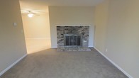 This single-story space centers around a living area defined by a standout stacked-stone fireplace, which serves as the room's focal point. The fireplace features a multi-toned stone surround with varying shades of gray, brown, and tan, complemented by a raised stone hearth and classic glass-paneled doors that add both character and functionality. A clean white mantel shelf crowns the surround, providing a simple yet elegant finishing touch.

The living area is finished with wall-to-wall neutral carpeting in a warm beige tone, contributing to a comfortable and understated aesthetic throughout. The walls are painted in a soft, warm beige that coordinates seamlessly with the flooring, creating a cohesive and inviting atmosphere. White baseboards frame the perimeter of the room, providing clean definition between the walls and floor.

A ceiling fan is visible in the adjacent area, suggesting the space flows into a connected room or dining area, offering both practicality and flexibility in layout. The overall design is straightforward and functional, with neutral finishes that provide a versatile backdrop suitable for a variety of interior styles and furnishings.