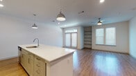 This single-floor unit offers a clean, contemporary design with an open-concept layout that seamlessly connects the kitchen and living areas. The kitchen is well-appointed with gray shaker-style cabinetry, white quartz countertops, a subway tile backsplash, and a full suite of stainless steel appliances, including a refrigerator, range, and microwave. A central kitchen island with an integrated sink and pendant lighting serves as a functional focal point, providing additional prep and seating space.

The living area is bright and spacious, benefiting from natural light through large windows and a set of double doors. A ceiling fan, recessed lighting, and built-in shelving add both practicality and character. Wood-look flooring extends throughout the main living spaces, creating a cohesive and warm aesthetic.

A dedicated entry area features wall-mounted hooks for convenient storage. The bedroom is carpeted for comfort, and an adjacent walk-in closet provides ample storage with dual hanging rods and shelving. The full bathroom includes a vanity with a granite-topped countertop, gray cabinetry, a tub-shower combination with white tile surround, and wood-look flooring.