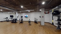 This fitness center offers a comprehensive workout environment housed within a single open floor plan. The space features a distinctive industrial aesthetic, with an exposed concrete ceiling accented by visible ductwork and recessed lighting that evenly illuminates the entire area. Light wood-look flooring spans the full length of the gym, providing a clean and durable surface for a wide range of exercise activities.

The cardio zone is well-appointed with multiple treadmills, elliptical trainers, air bikes, and stationary cycling bikes — several equipped with individual display screens for an engaging workout experience. A dedicated strength training area includes a multi-functional cable trainer and a full rack of dumbbells in graduated sizes, catering to both beginners and advanced users. Additional equipment such as a rowing machine, medicine balls, battle ropes, and balance trainers rounds out the functional fitness offerings.

Wall-mounted flat-screen televisions are positioned throughout the space, keeping members entertained during their sessions. A large mirrored wall in one section of the gym enhances the sense of openness and allows users to monitor their form. Storage racks keep accessories organized and accessible. The overall ambiance is practical, focused, and well-maintained, making it a fully functional facility suited for a diverse range of fitness routines.