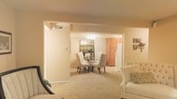 This single-floor residence offers a cohesive and comfortable living environment characterized by warm neutral tones and soft, plush carpeting throughout. The open-concept layout seamlessly connects the living room, dining area, and kitchen, creating a spacious and airy feel ideal for both everyday living and entertaining. The living room is furnished with classic seating and provides an easy flow into the dining space. The dining area serves as a natural focal point, featuring a round glass-top table surrounded by elegantly upholstered chairs and anchored by a large decorative mirror that reflects light and adds visual depth to the room. A ceiling fan overhead ensures comfort year-round. Adjacent to the dining area, the kitchen is outfitted with warm wood-toned cabinetry and includes full-size appliances, offering ample storage and functional workspace. The overall ambiance of the space is cozy and welcoming, with a consistent warm color palette tying each area together harmoniously. The single-level layout ensures easy accessibility and a practical flow between spaces, making this residence both functional and aesthetically pleasing.
