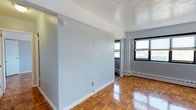 This single-floor space showcases a straightforward and functional interior design. The most notable flooring feature is the classic parquet hardwood, finished to a polished sheen that reflects light and adds warmth to the overall aesthetic. The walls are painted in a soft, neutral light blue-gray tone, contributing to a calm and understated ambiance throughout.

A dividing wall segments the interior, creating a sense of separation between areas while maintaining an open flow through a visible passageway. Natural light enters through a black-framed window, which also provides views to the exterior. Beneath the window sits a baseboard heating unit, a practical feature for climate control during cooler months.

White baseboards run along the perimeter of the space, providing a clean contrast against both the flooring and the wall color. A smoke detector is mounted on the wall, and a single electrical outlet is visible near the floor. The white ceiling is smooth and uniform, completing the clean, no-frills presentation of the space. Overall, the interior offers a well-maintained, move-in-ready environment with functional architectural elements throughout.