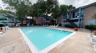 This residential community showcases a well-maintained outdoor amenity area centered around a generously sized rectangular swimming pool with clear, turquoise water. The pool deck is finished in a light-toned concrete surround, providing ample space for relaxation and movement. A selection of lounge chairs and a shaded patio table with a green umbrella are positioned poolside, offering residents comfortable spots to unwind. The pool area is enclosed by a black metal fence, ensuring a secure and defined space. Towering mature trees frame the surroundings, lending a canopy of natural shade and a lush, established character to the environment. The backdrop features classic multi-story brick buildings with exterior staircases and balconies, reflecting a traditional residential architectural style. The combination of the verdant tree coverage, brick facades, and inviting pool creates a community atmosphere that balances comfort with practicality. Signage is visible near the pool entrance, indicating community guidelines. Overall, the outdoor space presents a clean, functional, and welcoming environment suited for both leisure and community living.