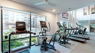 This modern fitness center offers a generously sized, single-floor layout designed to accommodate a full range of workout needs. The space is defined by its dramatic double-height ceilings in the main exercise area, which create an open, airy atmosphere and are complemented by large floor-to-ceiling windows that flood the interior with natural light and provide views of the surrounding outdoor environment. Structural columns punctuate the floor plan, subtly dividing the space into distinct workout zones without compromising the overall sense of openness. The cardio zone is well-appointed with treadmills, elliptical trainers, rowing machines, and stair climbers, many positioned to take advantage of the outdoor views. The strength training area features a robust selection of cable machines, multi-functional cable towers, free weights, dumbbells, weight benches, and a power rack, catering to a wide variety of training styles. Wall-mounted flat-screen televisions are positioned throughout for entertainment during workouts. Full-length mirrors along the walls enhance the sense of space and support proper form during exercise. Ceiling fans and recessed lighting provide comfort and visibility throughout. The flooring consists of durable, neutral-toned carpet tiles suited for high-traffic fitness use. The overall design balances a clean, contemporary aesthetic with a highly functional and motivating workout environment.