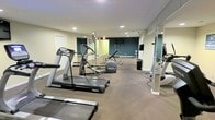 This fitness center is a dedicated, single-floor exercise facility designed to support a well-rounded workout routine. The space is generously sized, providing ample room to move freely between equipment and exercise zones without feeling cramped. Cardio equipment is a highlight of the room, with multiple Precor treadmills and an elliptical trainer positioned to give users a comfortable workout experience. A flat workout bench and a medicine ball cater to strength and functional training needs, while additional equipment is visible along the perimeter. Floor-to-ceiling mirrors line the walls on multiple sides, visually expanding the space and allowing users to monitor their form during exercise. The mirrored walls are complemented by accent wall painted in a deep teal tone, adding a modern, energetic touch to the otherwise neutral color palette of light beige walls and carpeted flooring. Recessed ceiling lighting provides bright, even illumination throughout the space. A designated area with a wall-mounted television offers an entertainment option during workouts. The overall ambiance is clean, functional, and professional, making this fitness center a practical and inviting space for daily exercise.