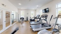 Located in Lewes, Delaware, this fitness center at 36916 Crooked Hammock Way offers a comprehensive exercise facility designed to support a wide range of workout routines. The space is finished with clean white walls, recessed lighting, and durable cork-style flooring that provides both comfort and resilience underfoot. Multiple white-framed windows line the walls, allowing ample natural light to brighten the interior and offer views of the surrounding greenery. A wall-mounted clock keeps workouts on schedule, while a flat-screen TV mounted near the cardio area adds entertainment options. The room is well-stocked with cardio equipment, including multiple treadmills, an elliptical trainer, and a recumbent exercise bike. A dedicated strength-training area features a weight bench and a tiered dumbbell rack. For functional fitness and flexibility training, the space includes a medicine ball rack with assorted weighted balls, yoga mats, and stability balls neatly organized on built-in open shelving. A large wall mirror visually expands the space and assists with form correction during workouts. Recessed ceiling lighting supplements the natural light, ensuring the room remains well-lit at all times. The overall ambiance is clean, organized, and functional, making this fitness center a practical and inviting amenity.