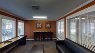 This game room in Conroe, Texas, is a lively, single-floor recreational space designed for entertainment and casual gatherings. The interior is immediately distinguished by its bold burnt-orange walls, accented by crisp white crown molding and white-trimmed doors and window casings, creating a warm and energetic atmosphere throughout.

The space is organized into two connected areas. The primary zone features a full-length shuffleboard table as a centerpiece, complemented by a wall-mounted flat-screen television flanked by a collection of framed artwork. A black leather sofa and a low coffee table provide comfortable lounge seating, making the area suitable for both active play and relaxed viewing. Flush-mounted fluorescent lighting illuminates the space evenly from above, while a textured ceiling adds architectural character.

The adjacent area includes a bar-height counter with seating for several guests, accompanied by additional framed artwork on the walls. Multiple large windows fitted with horizontal blinds allow natural light to filter in while maintaining privacy and offering views of the surrounding outdoor environment.

Patterned carpet tiles in muted earth tones cover the floors throughout, adding texture underfoot. A hallway connects the game room to additional areas of the broader facility, enhancing its accessibility. The overall design balances a recreational, social atmosphere with functional, well-appointed furnishings.