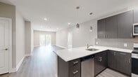 This well-appointed single-floor unit showcases a thoughtful, contemporary design throughout. The open-concept kitchen flows seamlessly into the living area, creating a bright and connected space ideal for everyday living. The kitchen is outfitted with dark wood-tone cabinetry, white quartz countertops, a tiled backsplash, and a full suite of stainless steel appliances including a gas range, over-the-range microwave, dishwasher, and a French door refrigerator. Pendant lighting over the peninsula island adds a refined finishing touch. Light-toned luxury vinyl plank flooring extends through the main living areas, complementing the neutral warm-gray wall palette throughout. The spacious living room benefits from a sliding glass door that opens to a private balcony, inviting natural light and outdoor views. Bedrooms are finished with plush carpeting and receive natural light through window treatments, offering a comfortable and quiet retreat. The primary bathroom features large-format tile flooring, a double-sink vanity with white countertops, and a glass-enclosed walk-in shower with decorative tile surround. A secondary bathroom includes a single vanity with matching dark cabinetry and contemporary fixtures. Recessed lighting, clean white trim, and modern hardware unify the design across the entire unit.