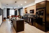 Located at 1912 S Jacaranda in Anaheim, CA, this single-floor residence showcases a striking contemporary design defined by a bold interplay of dark and light tones throughout. The open-concept layout seamlessly connects the kitchen and living areas, creating a spacious, cohesive feel. The kitchen is equipped with a full suite of black appliances, including a gas range, built-in microwave, refrigerator, and dishwasher, complemented by a dark center island with bar seating, a subway tile backsplash, and warm wood cabinetry with open shelving. An adjacent workspace with floating shelves adds functional versatility to the layout. The living area features a rich cognac leather sectional sofa, a foosball coffee table, and a statement dark accent wall with padded wall panels, anchored by a ceiling fan and floor-to-ceiling drapes that frame generous windows, allowing ample natural light. The bedroom offers a dramatic tufted leather headboard wall, a dedicated walk-in closet, and a ceiling fan, with direct access to the en-suite bathroom. The bathroom is finished with large-format dark tile surrounding the soaking tub, warm wood cabinetry, and distinctive geometric black wall accents. Recessed lighting throughout enhances the space's modern, curated ambiance.