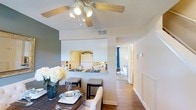 Located at 3300 Shannon Rd in Durham, North Carolina, this two-story residence offers a thoughtfully designed layout that balances comfort and functionality. The main living area features warm hardwood-style flooring that flows throughout, creating a cohesive and inviting atmosphere. The dining area is positioned beneath a classic ceiling fan with integrated lighting, providing both comfort and illumination. A dining table set with place settings and a fresh floral centerpiece anchors the space, lending it an elegant yet approachable feel. The kitchen connects seamlessly to the dining area through a half-wall pass-through, finished with white cabinetry and a clean, bright aesthetic. This open-concept arrangement enhances the sense of space and encourages natural interaction between the living and cooking areas. A hallway extending from the main living space leads to additional rooms, with glimpses of carpeted bedroom areas visible in the background, accented by natural light filtering through curtained windows. The neutral color palette — featuring soft whites, warm beiges, and cool gray accent walls — contributes to a calm and welcoming ambiance throughout. Spanning two floors, the home provides ample living space suited for a variety of lifestyles in the heart of Durham, NC.