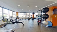 This fitness center offers a comprehensive workout environment across a single, open floor. The space is defined by large, wraparound windows that flood the interior with natural light and provide sweeping views of the surrounding urban skyline, creating an energizing atmosphere for exercise. Durable rubber flooring tiles cover the entire area, providing both safety and comfort underfoot.

The cardio zone is well-appointed with multiple treadmills, elliptical trainers, and stationary bikes, all positioned to take advantage of the expansive window views. A dedicated strength training section features a multi-station cable and weight machine, a full rack of dumbbells, adjustable benches, and an assortment of medicine balls and stability balls neatly organized on storage racks. Large wall-mounted mirrors in the weight area enhance the sense of space and allow users to monitor their form.

Motivational text is displayed on the walls, contributing to an encouraging workout environment. Multiple wall-mounted flat-screen TVs are positioned throughout for entertainment during workouts. The space also includes two private rooms accessible via gray doors, likely serving as changing areas or utility spaces. Recessed lighting in a drop ceiling ensures consistent illumination throughout. The overall design is modern, clean, and functional.