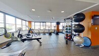 This fitness center offers a comprehensive workout environment across a single, open floor. The space is defined by large, wraparound windows that flood the interior with natural light and provide sweeping views of the surrounding urban skyline, creating an energizing atmosphere for exercise. Durable rubber flooring tiles cover the entire area, providing both safety and comfort underfoot.

The cardio zone is well-appointed with multiple treadmills, elliptical trainers, and stationary bikes, all positioned to take advantage of the expansive window views. A dedicated strength training section features a multi-station cable and weight machine, a full rack of dumbbells, adjustable benches, and an assortment of medicine balls and stability balls neatly organized on storage racks. Large wall-mounted mirrors in the weight area enhance the sense of space and allow users to monitor their form.

Motivational text is displayed on the walls, contributing to an encouraging workout environment. Multiple wall-mounted flat-screen TVs are positioned throughout for entertainment during workouts. The space also includes two private rooms accessible via gray doors, likely serving as changing areas or utility spaces. Recessed lighting in a drop ceiling ensures consistent illumination throughout. The overall design is modern, clean, and functional.