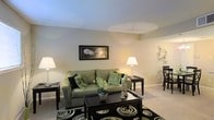 This single-floor residence offers a comfortable and thoughtfully arranged living environment. The living area serves as the heart of the space, characterized by warm, neutral-toned walls that create an inviting and relaxed ambiance. Soft wall-to-wall carpeting underfoot adds a sense of coziness throughout. The room is generously proportioned, providing ample space for seating and entertaining, with a layout that flows naturally into an adjacent dining area. A ceiling fan overhead contributes to the room's practical comfort. The living space features subtle architectural detailing, including a gently contoured ceiling that adds visual interest without overwhelming the room's understated elegance. Large, warm lighting from floor and table lamps enhances the welcoming atmosphere, compensating for the interior placement of the space. The open connection between the living and dining areas promotes a sense of continuity and ease of movement. Overall, the space strikes a balance between functional design and comfortable living, making it well-suited for both everyday relaxation and casual entertaining. The neutral palette and soft finishes throughout provide a versatile backdrop that accommodates a wide range of furnishing and décor styles.