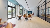 Located at 501 NW Seventh Ave in Fort Lauderdale, FL, this single-floor amenity space offers a generously sized, open-plan layout designed for socializing, relaxing, and recreation. The interior is defined by a contemporary aesthetic that blends clean architectural lines with warm, eclectic furnishings. Light hardwood flooring runs throughout, complemented by vibrant, layered Persian-style area rugs that anchor distinct seating zones.

Dramatic floor-to-ceiling windows and glass doors line the perimeter, flooding the space with natural light while framing lush views of tropical palms, landscaped gardens, and a pool area beyond. Bold navy blue modular sofas, cognac leather lounge chairs, and bistro-style café tables create a variety of comfortable gathering spots suited to different activities and group sizes.

Striking steel-framed glass partition walls divide the space into semi-private rooms, including what appears to be a game room featuring a foosball table and colorful bowling balls set against an exposed brick accent wall. A library nook with built-in illuminated shelving displays books and art objects, providing a quieter retreat. Recessed lighting, teal ceiling accents, and curated artwork contribute to a polished yet inviting atmosphere throughout this well-appointed amenity floor.