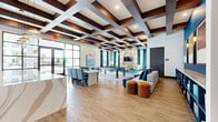 This single-floor amenity space offers a generously sized, open-plan layout designed for social gatherings and relaxation. The architectural highlight is a striking coffered ceiling with dark wood beams arranged in a grid pattern, adding depth and visual interest throughout the space. Wood-look tile flooring runs continuously underfoot, complementing the warm tones of the ceiling.

The main lounge area is divided into distinct functional zones. A seating area features mid-century-inspired striped armchairs arranged around a wooden coffee table, anchored by a vivid teal patterned area rug. A focal arched wood-paneled wall houses a mounted flat-screen television, flanked by floor-to-ceiling teal drapery that frames large windows allowing abundant natural light and outdoor views. A billiards table is centrally positioned within the open floor plan, while a dining table with upholstered chairs provides additional gathering space.

The kitchen area is well-appointed with quartz countertops, white cabinetry, hexagonal tile backsplash, open wood shelving, and stainless steel appliances. Large glass-panel entry doors connect to the exterior, further enhancing the bright, welcoming ambiance of the space.