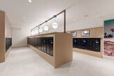 This well-appointed mail room combines practical functionality with refined design across a spacious, single-floor layout. At its center, a striking freestanding island of warm light wood cabinetry houses rows of sleek black individual mailboxes, elegantly illuminated by a series of globe pendant lights suspended from a bronze-toned overhead rail. Matching wall-mounted mailbox banks line the perimeter, ensuring ample mail storage capacity for a large residential community. A dedicated package retrieval area occupies a separate section of the room, where floor-to-ceiling smart parcel lockers finished in a deep gradient of teal to rose are arranged along two walls, accompanied by a digital touchscreen kiosk for seamless package pickup. The room is grounded by large-format light stone tile flooring that flows continuously throughout, lending a clean and cohesive aesthetic. Recessed ceiling lighting provides bright, even illumination across the space. A branded entry door featuring a bold gold monogram, flanked by angular wall sconces, serves as a distinctive focal point at the entrance. A built-in credenza with a mounted display screen and textured tile backsplash adds a further layer of sophistication. Curated artwork on the walls completes the upscale, modern ambiance of this thoughtfully designed communal space.