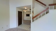 This two-story residence greets visitors with a tiled entryway that transitions smoothly into carpeted living areas beyond. The entry foyer is defined by clean white walls and a dark-toned front door, creating a welcoming contrast. Flush-mount ceiling light fixtures illuminate the space with a soft, consistent glow throughout the main level.

From the entryway, the layout opens toward an adjoining living area finished in wall-to-wall carpet, lending a comfortable, quiet feel underfoot. A pass-through opening or window is visible toward the rear, suggesting a connected kitchen or dining area with cabinetry visible in the background.

A staircase with white-painted balusters and a warm wood handrail rises prominently along one side of the entry, providing access to the second floor. The staircase design is classic and functional, with open shelving-style detailing on the side panel adding a subtle architectural touch.

Throughout the space, the neutral white walls and light-colored finishes create a bright, clean atmosphere that offers flexibility for various interior styles. The overall layout emphasizes practicality, with clearly defined transition zones between the entry, living, and upper-level spaces.