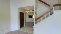 This two-story residence greets visitors with a tiled entryway that transitions smoothly into carpeted living areas beyond. The entry foyer is defined by clean white walls and a dark-toned front door, creating a welcoming contrast. Flush-mount ceiling light fixtures illuminate the space with a soft, consistent glow throughout the main level.

From the entryway, the layout opens toward an adjoining living area finished in wall-to-wall carpet, lending a comfortable, quiet feel underfoot. A pass-through opening or window is visible toward the rear, suggesting a connected kitchen or dining area with cabinetry visible in the background.

A staircase with white-painted balusters and a warm wood handrail rises prominently along one side of the entry, providing access to the second floor. The staircase design is classic and functional, with open shelving-style detailing on the side panel adding a subtle architectural touch.

Throughout the space, the neutral white walls and light-colored finishes create a bright, clean atmosphere that offers flexibility for various interior styles. The overall layout emphasizes practicality, with clearly defined transition zones between the entry, living, and upper-level spaces.