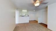 This single-floor unit presents an open and functional layout that integrates the living area and kitchen in a cohesive design. The living space is finished with wall-to-wall carpet in a neutral beige tone, creating a warm and comfortable atmosphere. A ceiling fan with an integrated light fixture is centrally positioned overhead, providing both airflow and ambient lighting to the room.

The kitchen is defined by a half-wall pass-through counter that opens toward the living area, maintaining a sense of openness while providing a dedicated workspace. White cabinetry lines the upper portion of the kitchen walls, complemented by white appliances including a microwave and refrigerator. Under-cabinet fluorescent lighting brightens the kitchen workspace.

Transitioning toward the back of the unit, the flooring shifts to a hardwood-style surface, adding visual contrast. A set of bifold closet doors offers generous storage, while additional doors lead to separate rooms or utility areas. The walls throughout are finished in a clean white, contributing to a bright and neutral palette. The overall design is straightforward and practical, emphasizing ease of use and livability.