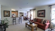 This single-floor residence offers an open-concept floor plan that integrates the kitchen, dining, and living areas into one cohesive, flowing space. The kitchen features warm wood cabinetry, light countertops, and a raised breakfast bar with barstool seating — ideal for casual dining or entertaining. Appliances are conveniently built in, and the layout allows for easy interaction between the kitchen and the rest of the living space. Adjacent to the kitchen, a dedicated dining area accommodates a full table and chairs, bathed in natural light from nearby windows dressed with decorative curtains. The living room is generously proportioned, anchored by a rustic wooden coffee table and a comfortable sofa with decorative pillows. Framed wall art and a coordinated floor lamp add warmth and character to the space. Light-toned carpeting runs throughout, contributing to the home's soft and inviting ambiance. Neutral wall tones and a textured ceiling complete the classic residential aesthetic. Natural light filters through multiple windows, enhancing the open and airy feel of the entire main living area.