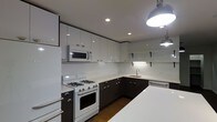 This single-floor space makes a striking impression with its thoughtfully designed kitchen as a central highlight. The kitchen showcases a bold two-tone design, pairing crisp white upper cabinetry with deep espresso-toned lower cabinets, creating a sharp and contemporary contrast. White quartz countertops run throughout, providing ample prep space and a clean, polished aesthetic. A classic white subway tile backsplash ties the design together with a timeless touch. The kitchen is equipped with a white gas range and oven, an over-the-range microwave, and a stainless steel sink set beneath open shelving brackets that offer additional storage or display options. Recessed ceiling lighting is complemented by an industrial-style pendant light, casting warm, focused illumination across the workspace. The hardwood flooring adds a natural warmth that grounds the otherwise sleek, monochromatic palette. The layout is designed for efficient workflow, with generous counter space and well-organized cabinetry providing abundant storage. The overall ambiance is clean, modern, and functional — a space that balances style with practicality in equal measure.