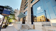Located in the heart of the city, 735 West Apartments is a modern high-rise residential building with a striking stone and glass exterior façade. The building's entrance features a covered canopy, stairway access, and large street-level windows that give it a polished, urban presence among the surrounding downtown skyline.

The featured amenity space is a generously sized fitness center designed to support a full range of workout routines. The gym is finished with durable rubber flooring and benefits from high, exposed ceilings fitted with bright overhead lighting and visible ductwork, lending the space an open, industrial character. Large windows along the exterior wall invite natural light and provide outdoor views, enhancing the energizing atmosphere.

The fitness center is well-appointed with a variety of equipment, including True-brand treadmills and cardio machines positioned in front of wall-mounted flat-screen televisions for an engaging workout experience. The open floor area accommodates rowing machines, weight benches, and multi-use cable strength equipment. A full-length mirrored wall on one side of the room expands the sense of space and supports strength training activities. Connecting corridors with clean, neutral finishes provide seamless access throughout the amenity level. Overall, the fitness center reflects a functional and well-maintained environment suited for residents seeking convenient, on-site wellness facilities.