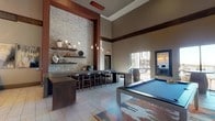 This single-floor amenity space is designed with a contemporary aesthetic that blends natural materials with modern finishes. Soaring ceilings create an expansive, airy atmosphere throughout, enhanced by large windows that draw in natural light and offer views of the surrounding exterior. Light-toned tile flooring runs continuously across the space, complementing the warm wood and stone accents found throughout.

The main lounge area centers around a pool table set atop a colorful abstract area rug, flanked by a floor-to-ceiling dark wood accent wall with vertical linear lighting and a wall-mounted television. A high-top bar counter with bar-height seating sits adjacent to a dramatic textured stone feature wall adorned with floating wood shelves and pendant lighting, creating an inviting social hub.

A dedicated business center offers built-in workstations equipped with desktop computers and a printer, lit by large rectangular pendant fixtures against the same stone backdrop. A seating nook features a live-edge wood console table beneath a gallery of large wood-slice wall art, providing a relaxed retreat. Additional lounge seating near tall windows offers a quiet, light-filled corner. The overall ambiance is warm, sophisticated, and thoughtfully curated with nature-inspired artistic elements.
